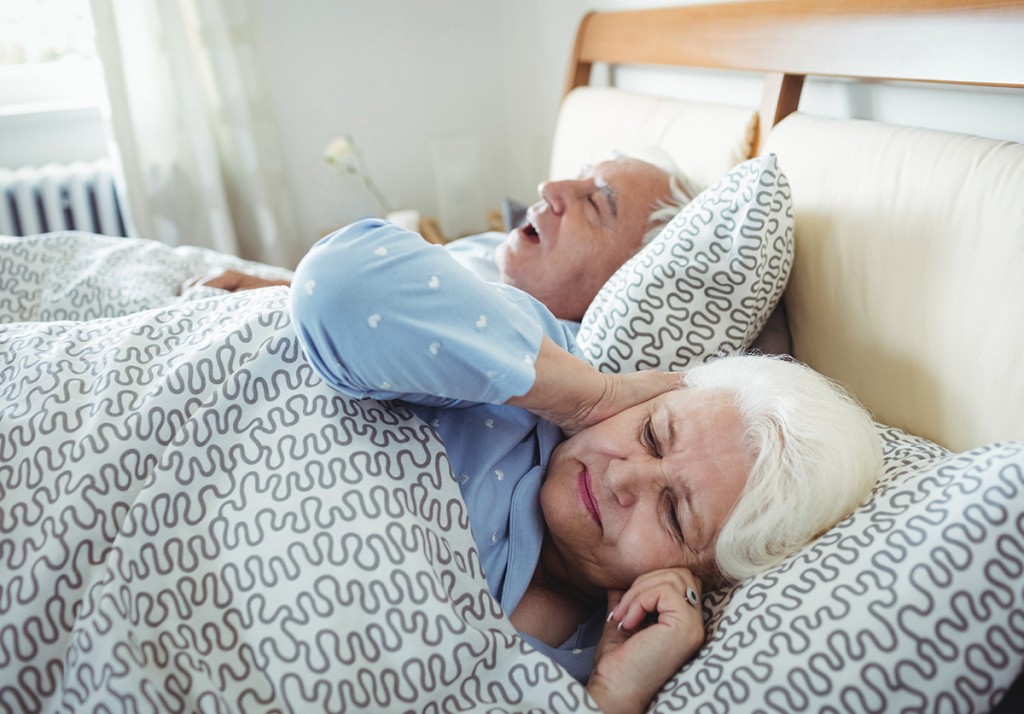 Older woman lying awake with her hand on her neck beside a sleeping man with his mouth open