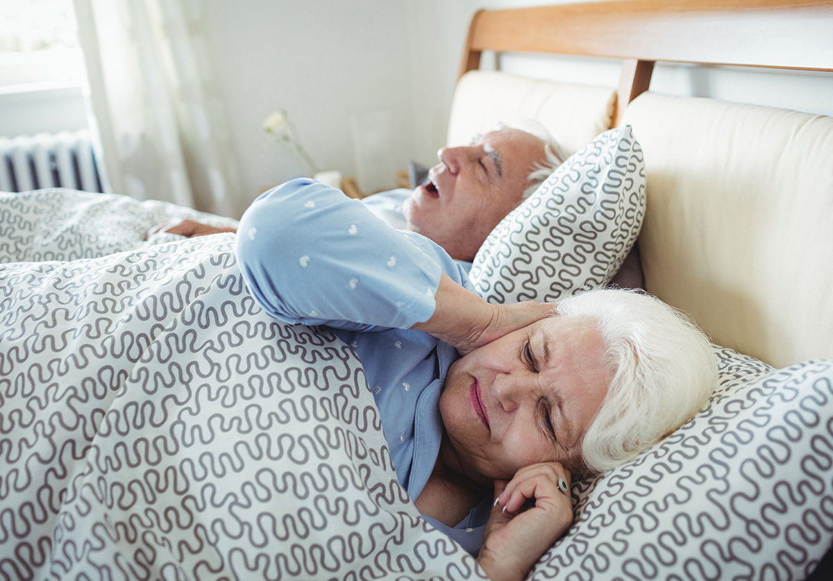 Older woman lying awake with her hand on her neck beside a sleeping man with his mouth open