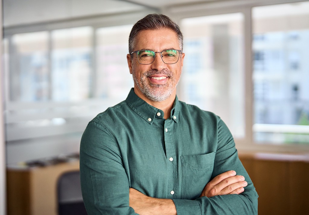 Smiling man with glasses and a green shirt standing with his arms crossed in an office