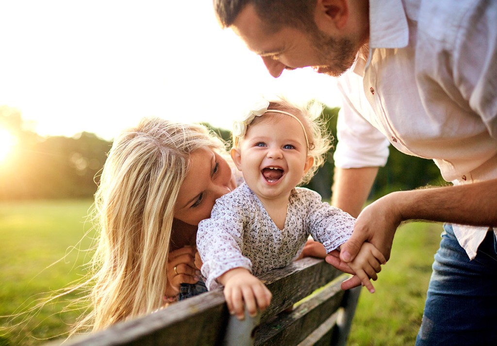 Smiling baby sitting between her parents outdoors while her father holds her hand