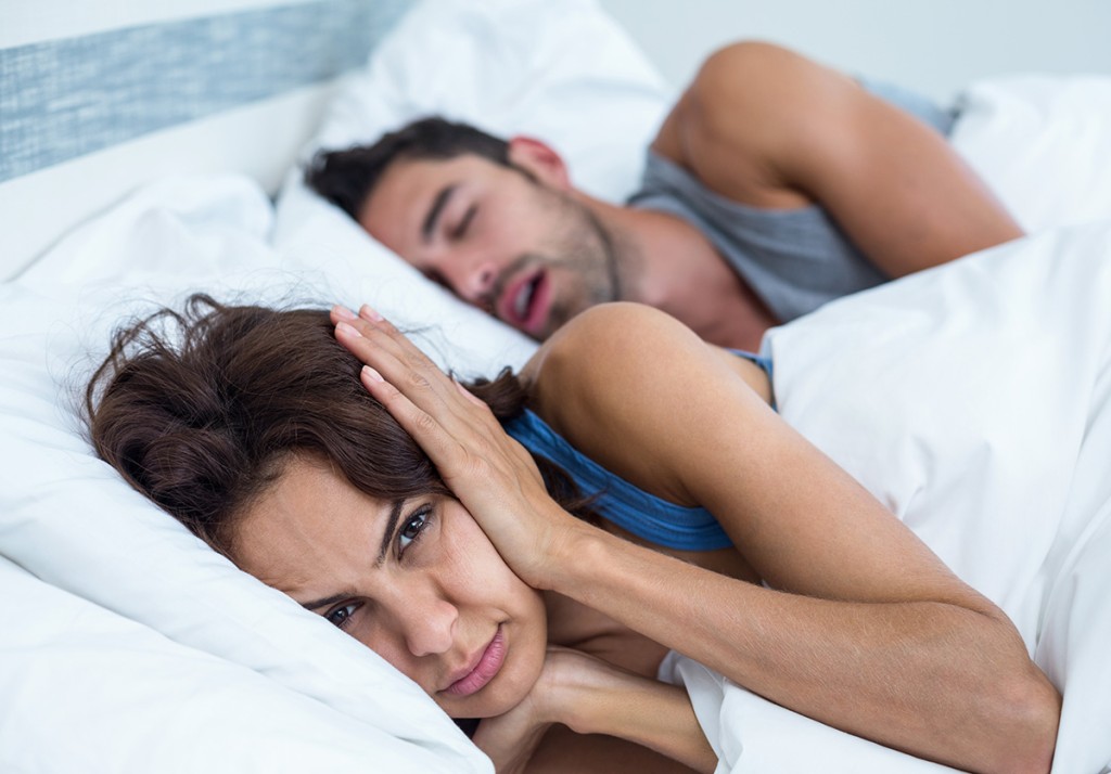 Woman covering one ear in bed while her partner sleeps with his mouth open