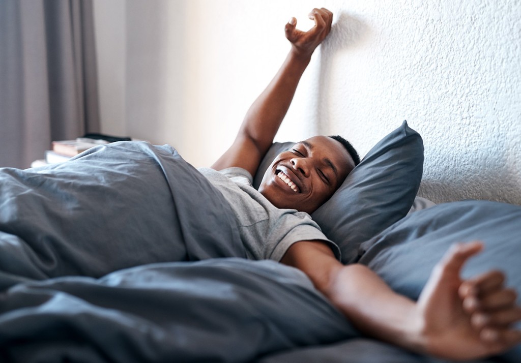 Smiling man stretching his arms while lying in bed under a gray comforter